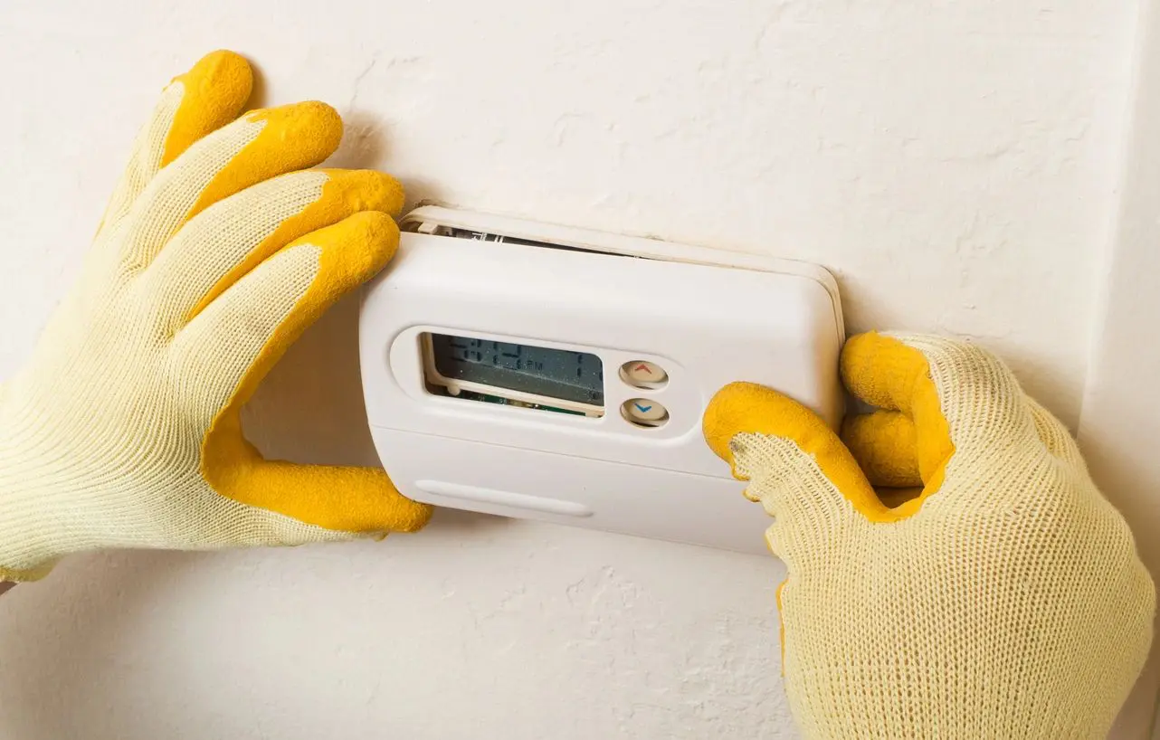 A person wearing yellow gloves is adjusting the temperature on an electric thermostat.